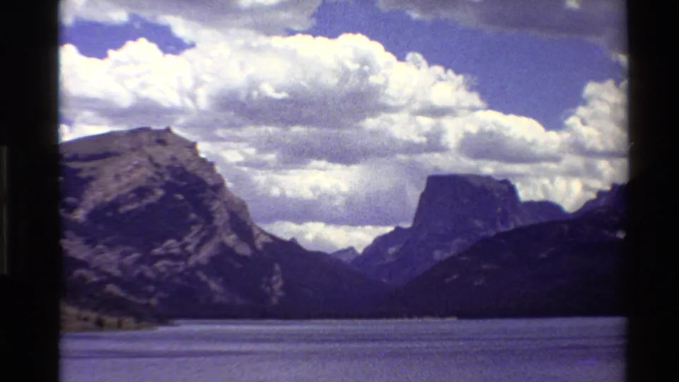 1980 Western USA 8mm frame showing a lake foreground, rocky mountain slope, distant peak, and billowing clouds across the sky.