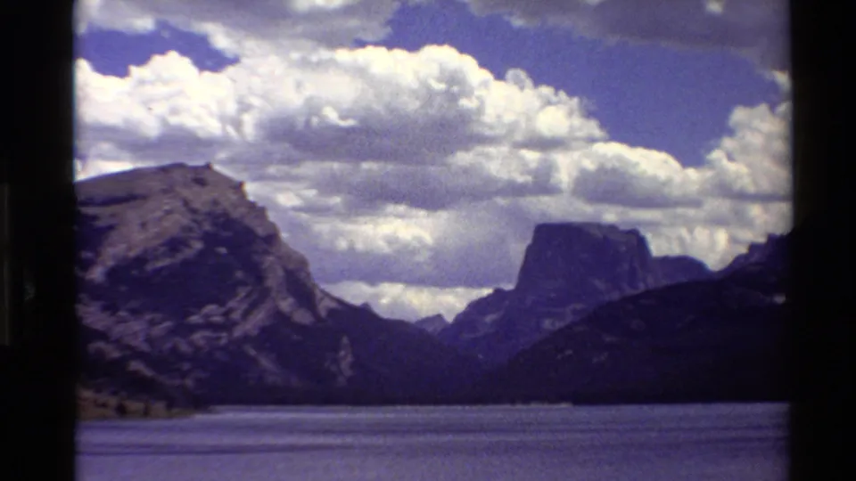 1980 Western USA 8mm frame with calm lake water, dark ridges, a flat-topped mountain peak, and bright cumulus clouds.