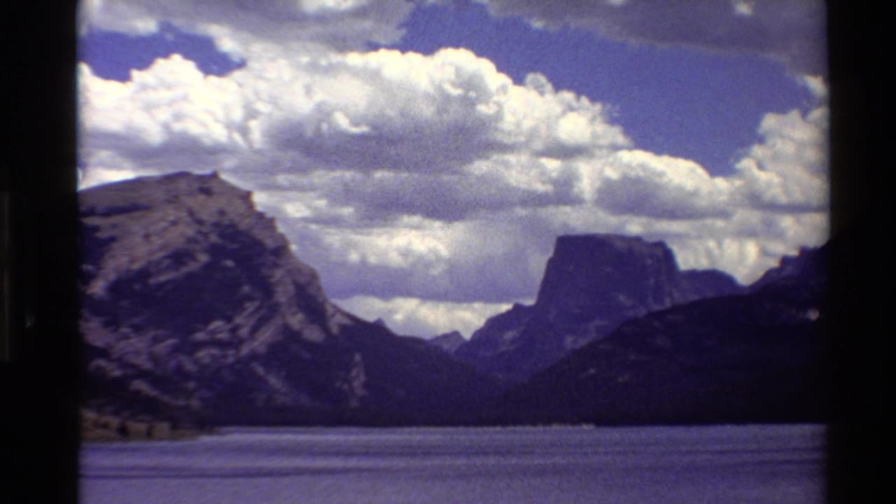 1980 Western U.S. 8mm frame of a calm lake, rugged mountain slopes, a flat-topped peak, and bright clouds under blue sky.