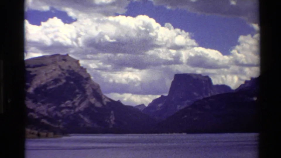 1980 Western U.S. 8mm frame of a calm lake, rugged mountain slopes, a flat-topped peak, and bright clouds under blue sky.