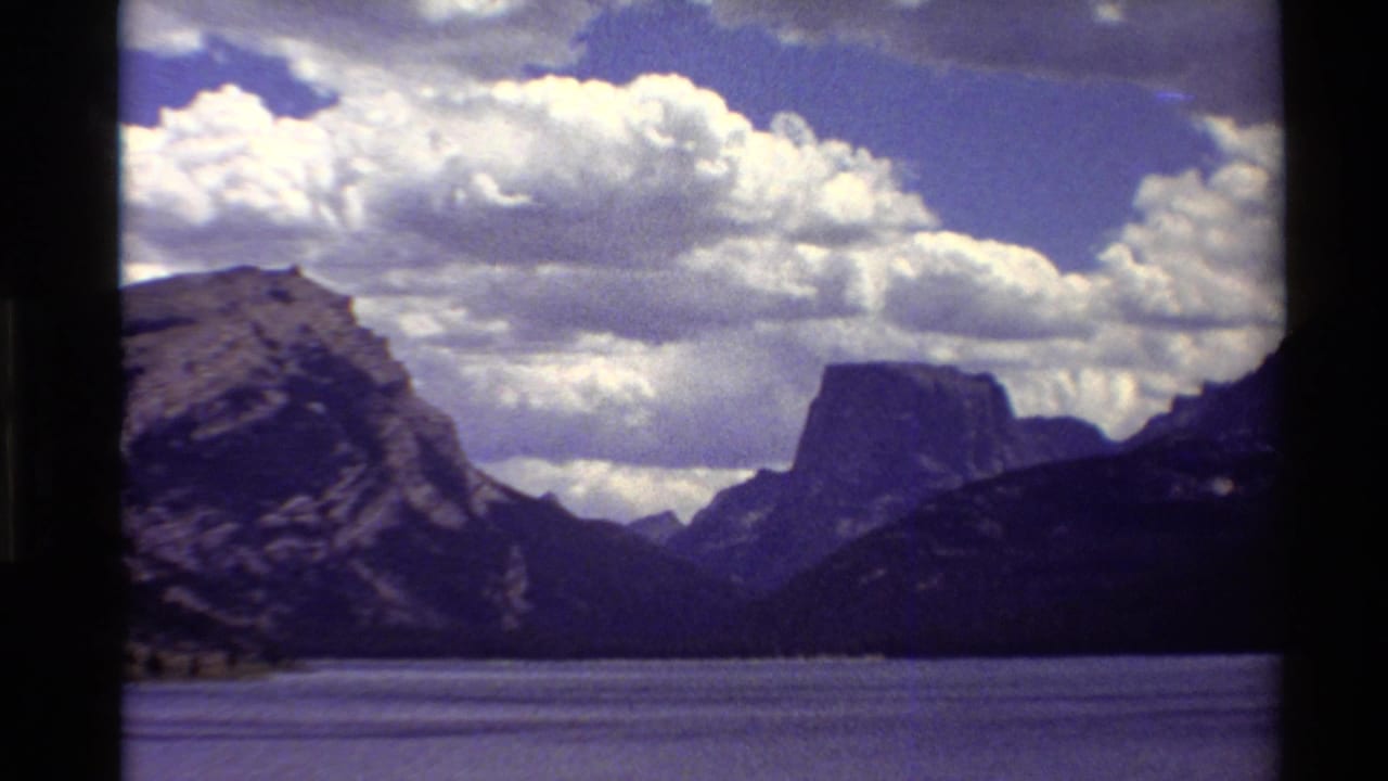 1980 Western U.S. 8mm frame showing a lake foreground, dark mountain ridges, rocky peaks, and dense white clouds.
