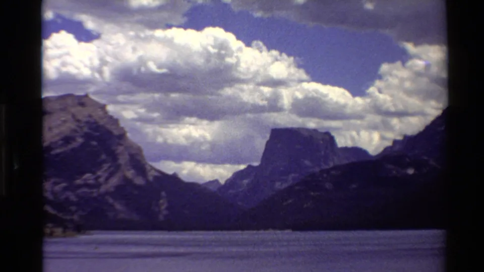 1980 Western U.S. 8mm frame with lake water, shadowed mountains, a central flat-topped peak, and billowing clouds.