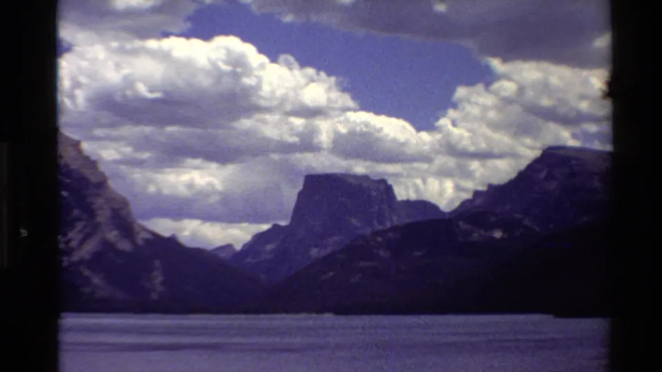 1980 Western U.S. 8mm frame of a lake, silhouetted ridges, rocky mountain forms, and bright cloud cover in a blue sky.