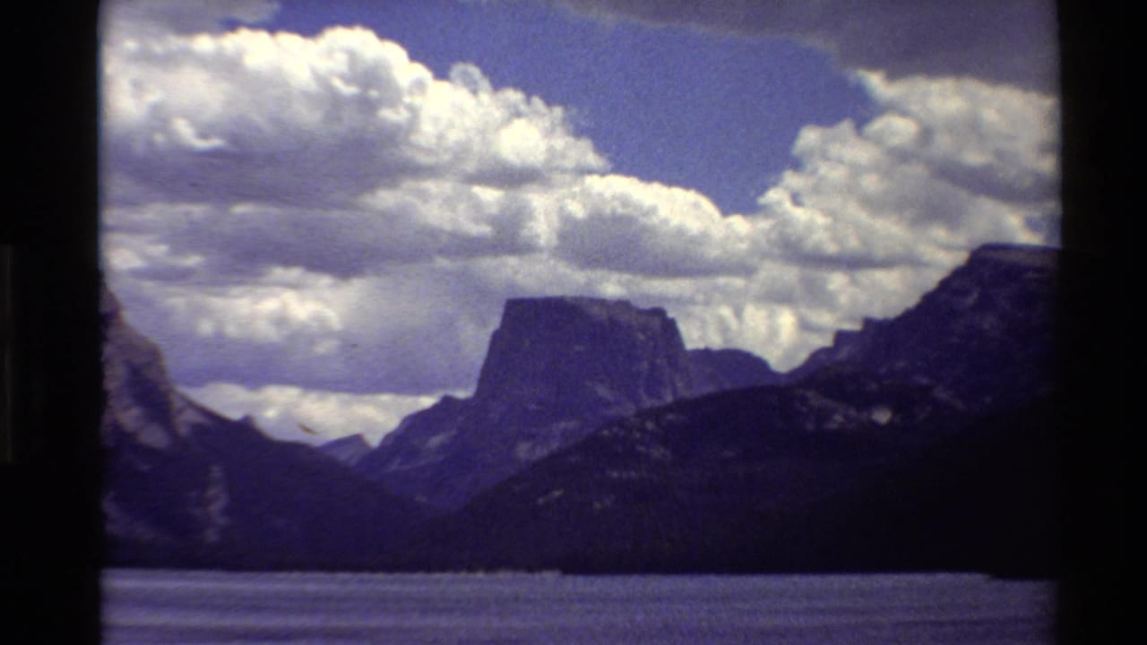 Wide lake view with dark mountain slopes, flat-topped peak, blue sky, and white clouds.