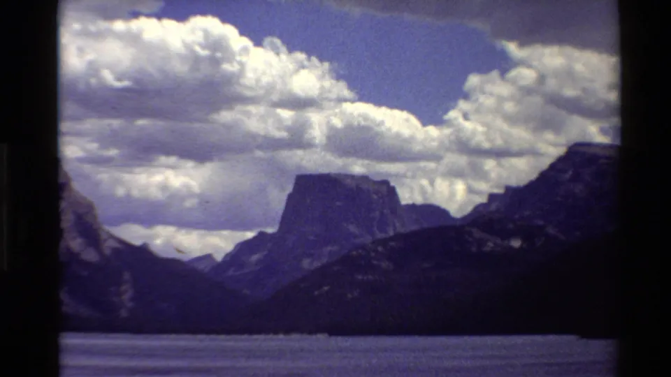 1980 Western USA 8mm frame showing a lake below dark mountain slopes and a distant flat-topped rocky peak under bright clouds.