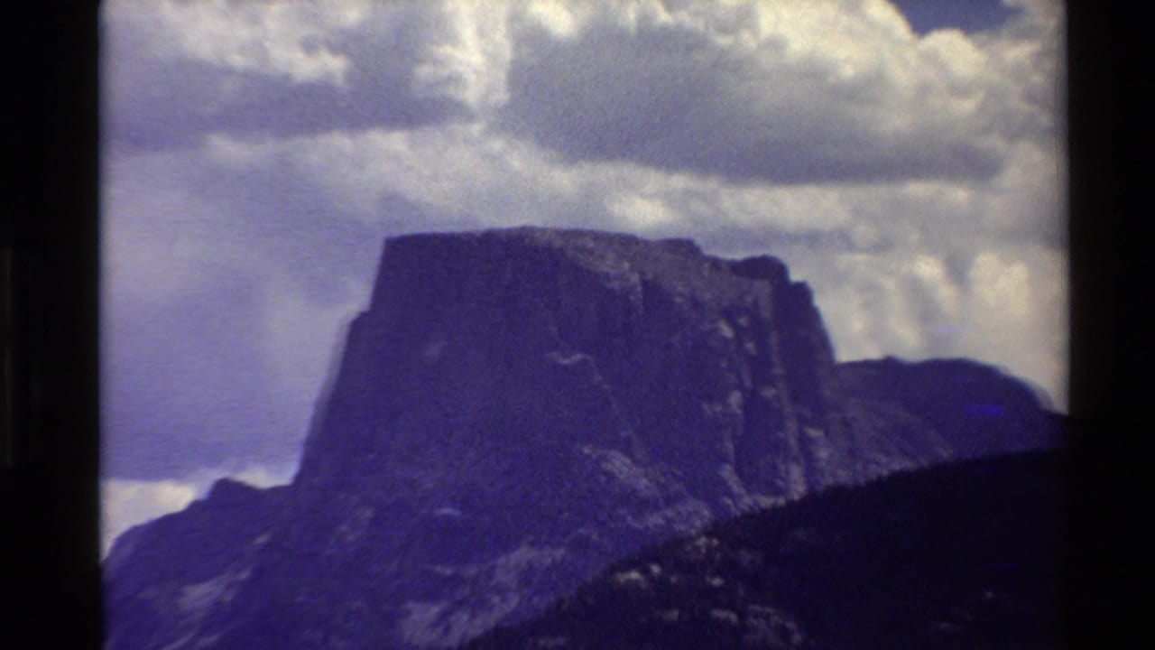 Closer 8mm view of a prominent flat-topped mountain in the Western USA, with dark slopes and puffy clouds overhead.