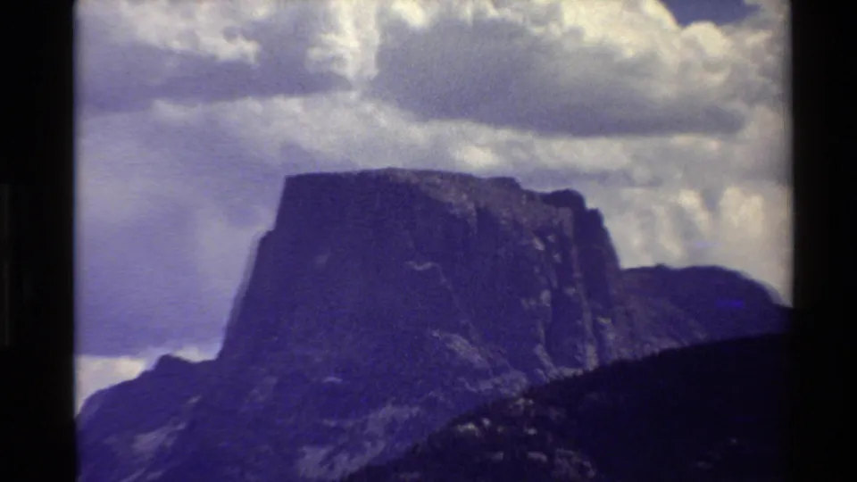 Closer 8mm view of a prominent flat-topped mountain in the Western USA, with dark slopes and puffy clouds overhead.