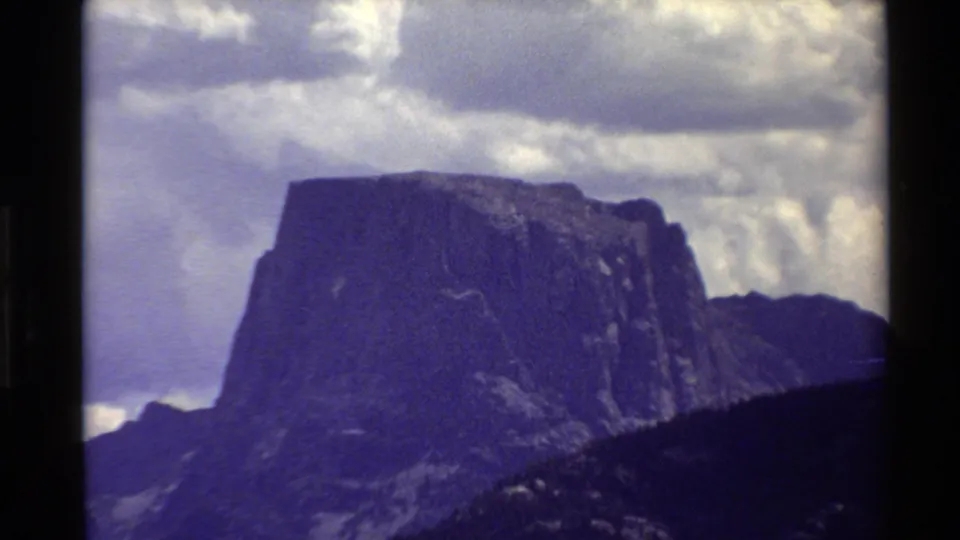 1980 8mm frame of a broad rocky peak filling the view, with dark lower slopes and white clouds behind it.