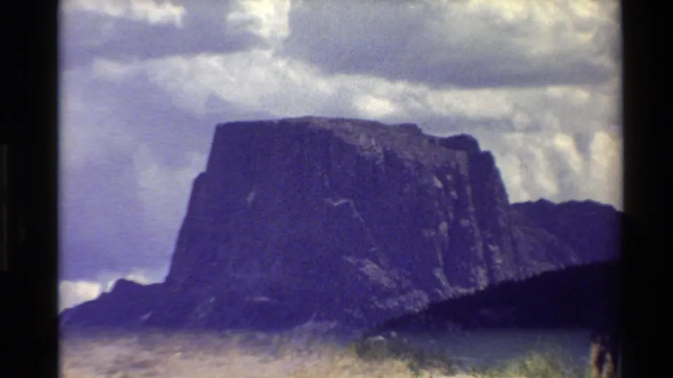 Western USA home-movie frame from 1980 showing a flat-topped rocky peak, shadowed slopes, and a bright clouded sky.