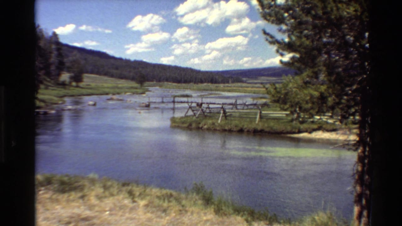 1980 Western U.S. 8mm view of a calm river valley with wooden rail fencing, green banks, pine hills, and a bright clouded sky.