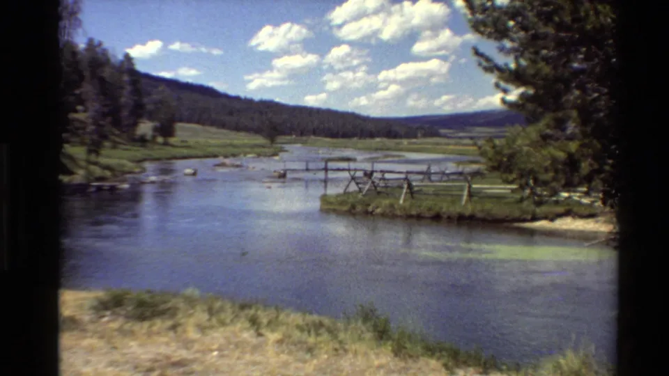 Western U.S. home-movie frame showing a broad river, rustic fencing near the bank, green hills, trees, and scattered white clouds.