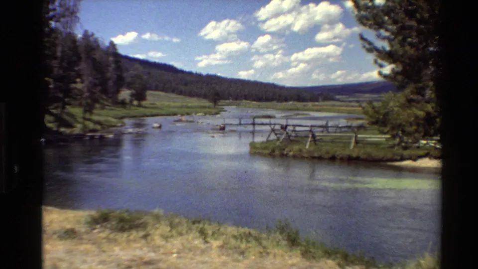 8mm travel view of a calm river and grassy bank with wooden fencing, a dark tree line, rolling hills, and a bright sky overhead.