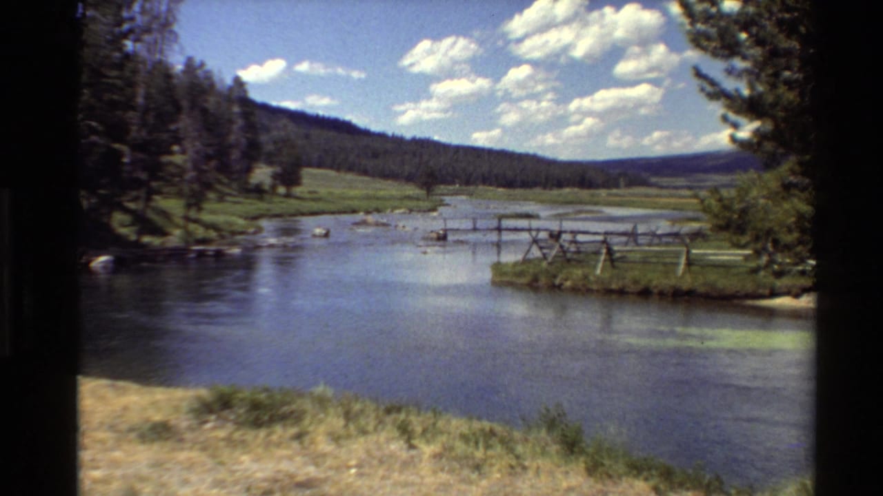 1980s 8mm view of a calm river, meadow banks, trees, distant hills, and scattered clouds in the Western United States.