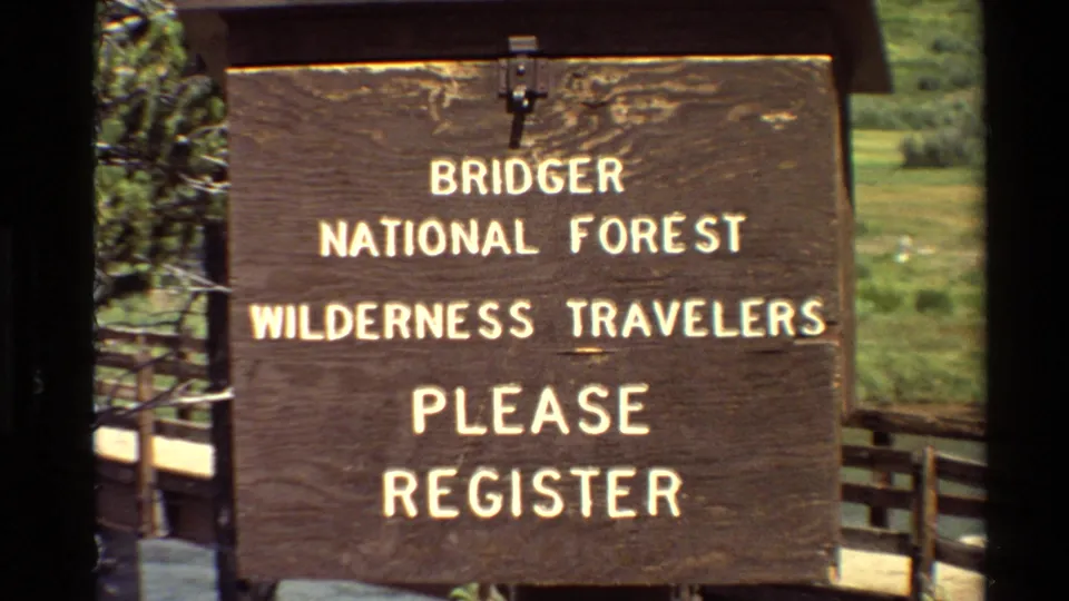 Close 8mm frame of a brown wooden Bridger National Forest registration sign with fence rails and green landscape behind it.