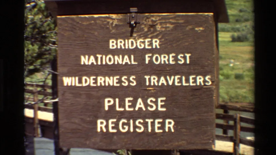 Final 1980s 8mm frame centers the Bridger National Forest wilderness traveler registration sign with fence and trees in the background.
