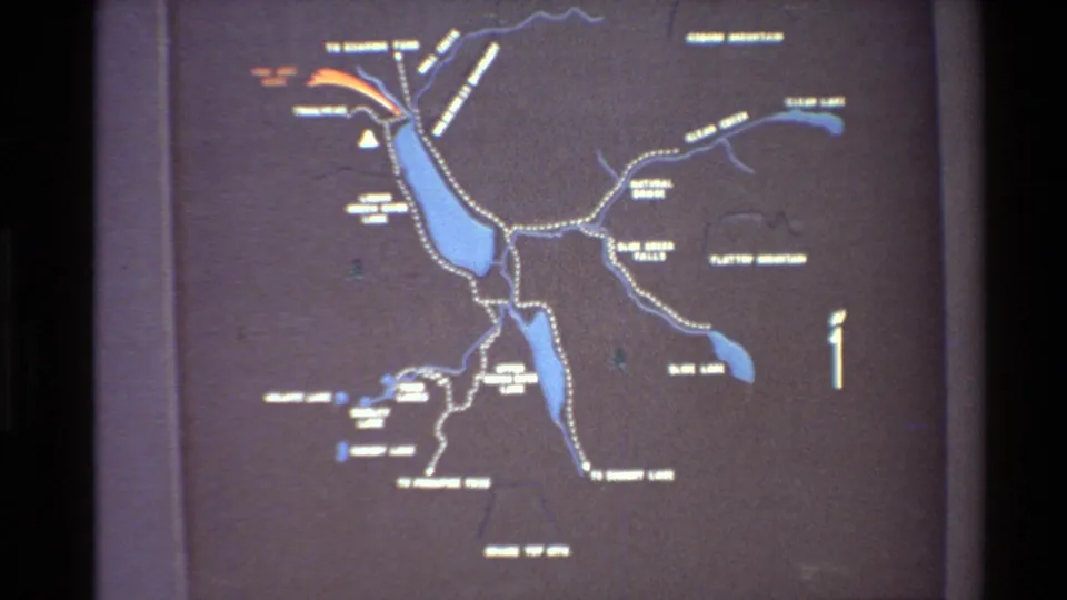 1980s home-movie frame with a broader view of a Western USA trail map, including lakes, routes, and white lettering.