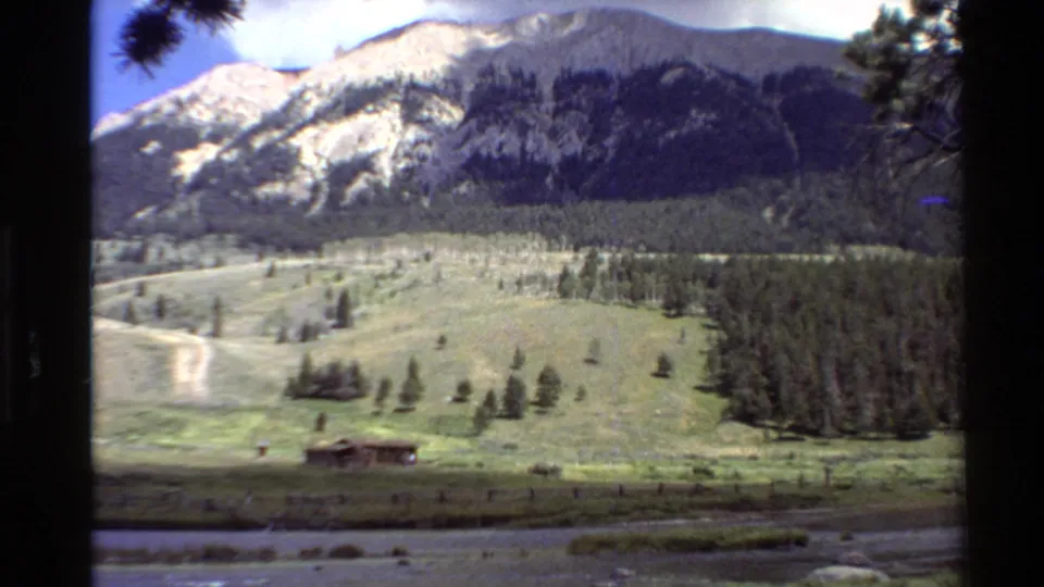Wide 1980 8mm view of a Western United States mountain valley with meadow, river, conifers, and a small cabin.