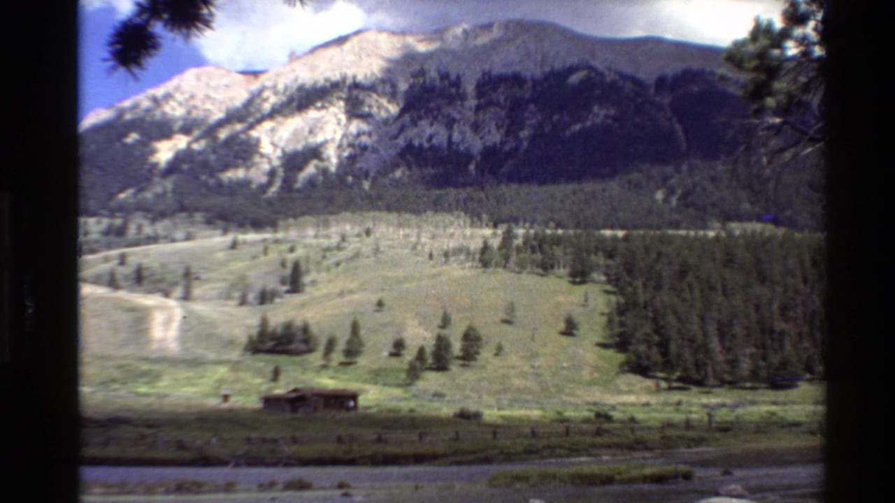 1980 Western U.S. 8mm view of a grassy mountain valley with scattered pines, a small cabin, a dirt path, and rocky high peaks.