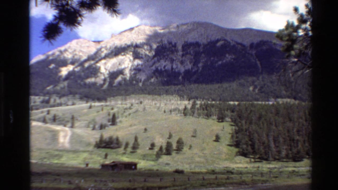 Wide valley landscape with a dirt road, cabin, pine forest, and mountain ridge.
