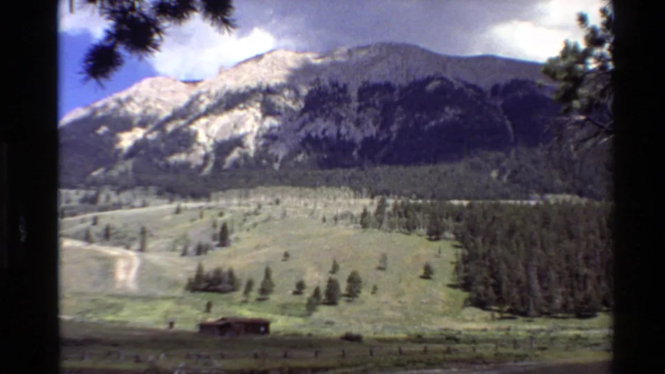Wide 8mm frame of a Western U.S. mountain valley in 1980, showing a dirt path, cabin, pine forest, and high ridge under clouds.