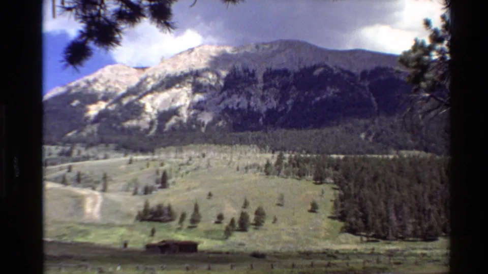 Home-movie view of a pine-framed Western U.S. valley, with a cabin in the meadow, forest beyond, and rocky mountain peaks.