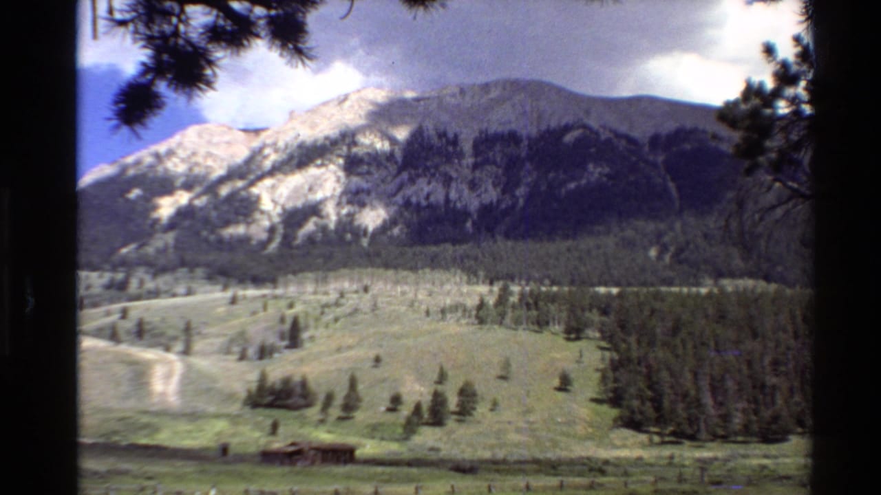 1980 Western U.S. 8mm frame showing a green mountain valley, scattered pines, a small distant structure, and clouded rocky slopes.