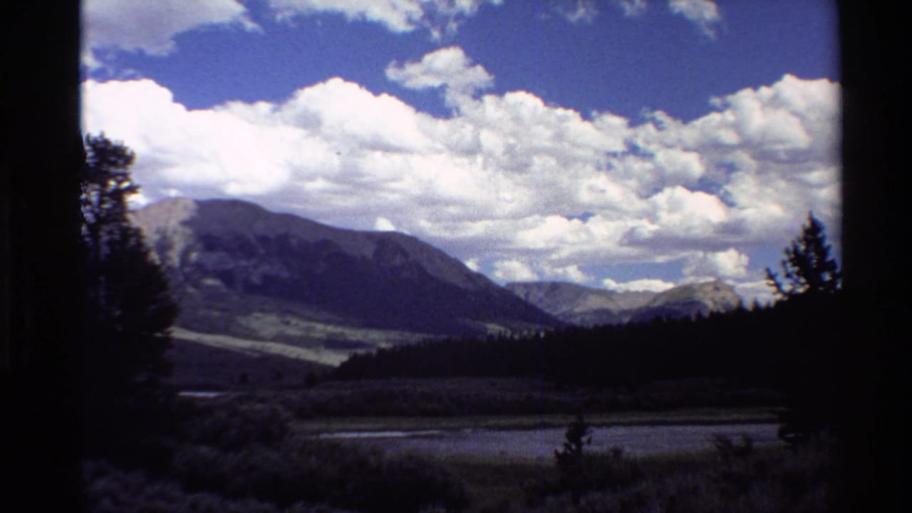 Shadowed valley and forest with mountains beneath a bright cloud-filled sky.