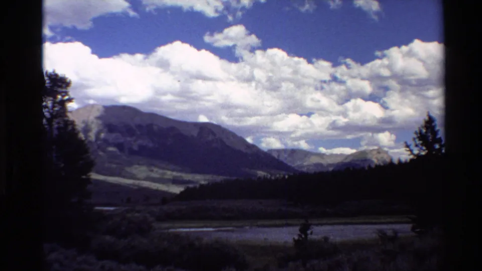 1980 Western U.S. 8mm frame looking across a shadowed valley, dark pine forest, distant mountains, and bright clouds.