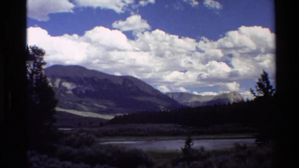 1980 Western U.S. 8mm frame showing a mountain valley, dark conifers, low water or wetland, and large cloud banks.