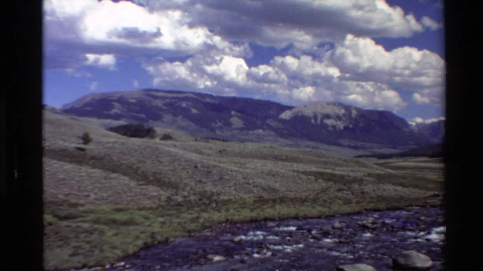 1980 Western U.S. 8mm frame of a shallow rocky stream, open valley terrain, distant mountain slopes, and cumulus clouds.