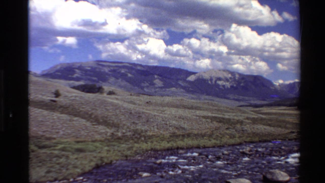 1980 Western U.S. 8mm frame showing a rocky stream in the foreground, open valley grassland, distant mountains, and bright cumulus clouds.