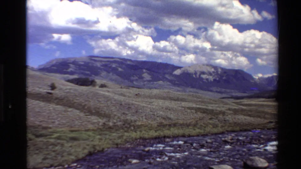 1980 Western U.S. 8mm frame showing a rocky stream in the foreground, open valley grassland, distant mountains, and bright cumulus clouds.