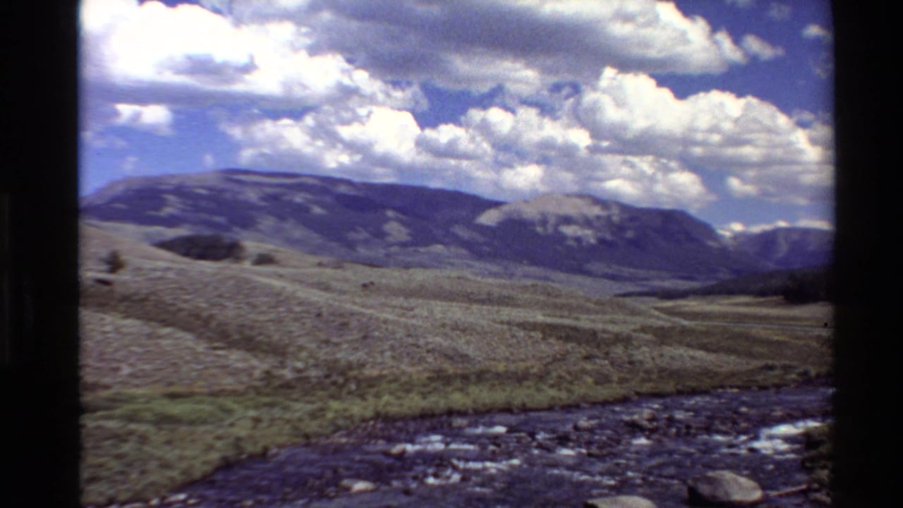 Open valley and stream with distant mountain ridges under a bright cloudy sky.