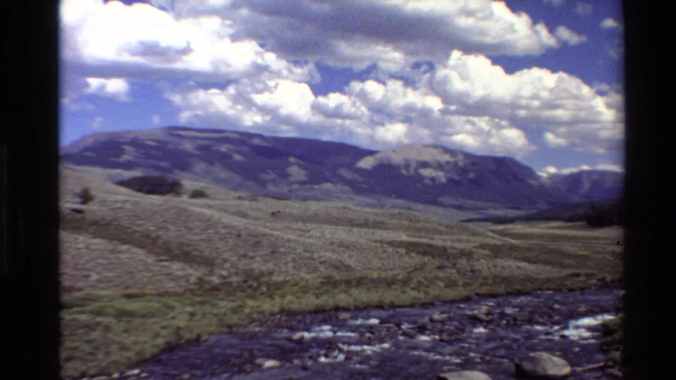 1980 Western U.S. 8mm frame with stream water in the foreground, low valley vegetation, mountain ridges, and sunlit clouds.