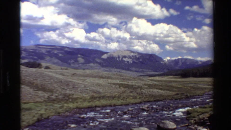 1980 Western U.S. 8mm frame showing a moving view past a stream toward rolling hills, distant mountains, and a partly cloudy sky.