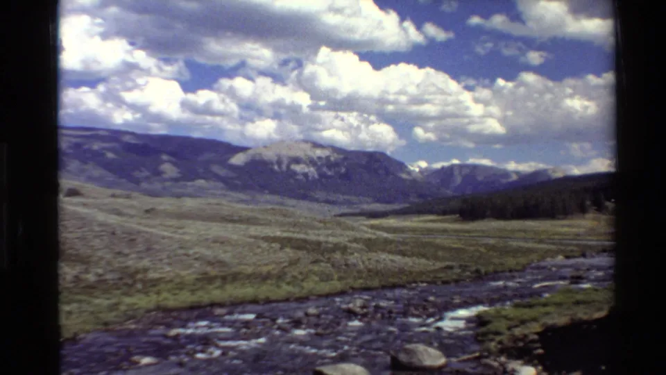 1980 Western U.S. 8mm frame with a rocky stream, open valley, distant mountains, heavy clouds, and a dark tree line at the right edge.