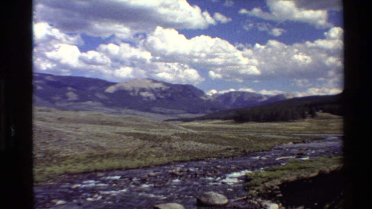 1980 8mm Western USA travel footage showing a rocky river, open grass plain, distant mountains, and bright cloud-filled sky.