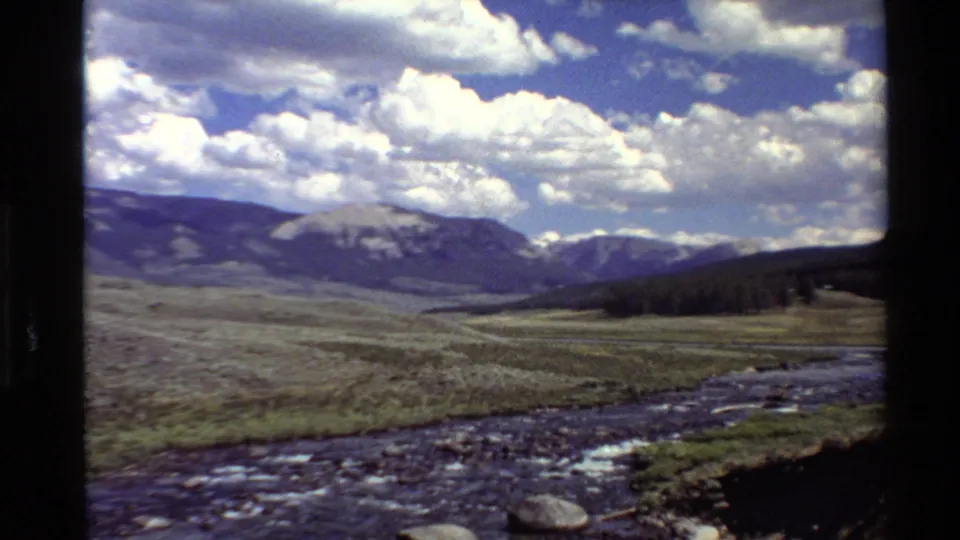 1980 8mm Western USA travel footage showing a rocky river, open grass plain, distant mountains, and bright cloud-filled sky.