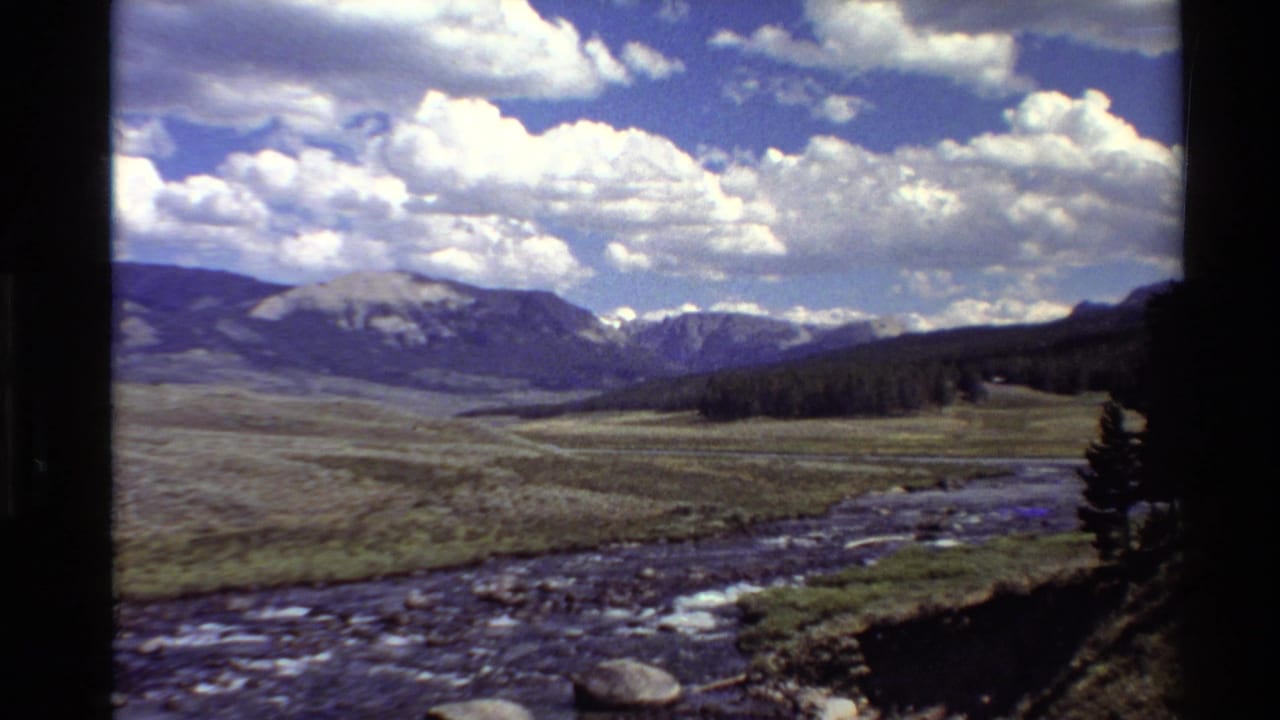 Mountain valley landscape with river, grassland, forest edge, and bright clouds.
