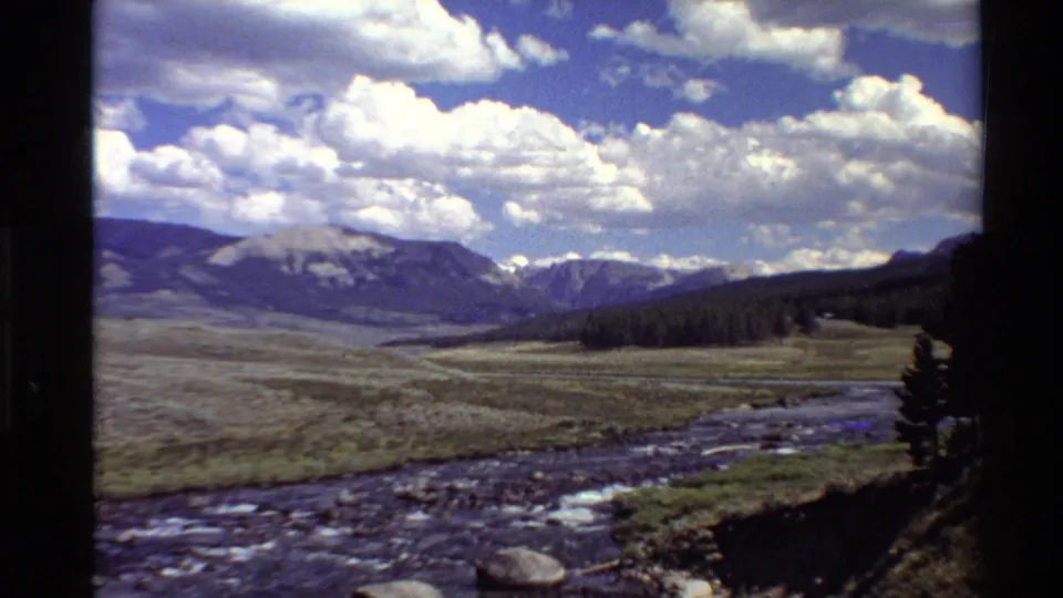 Western USA 8mm landscape view with a river in the foreground, broad meadow, forested slope, distant mountains, and high cumulus clouds.