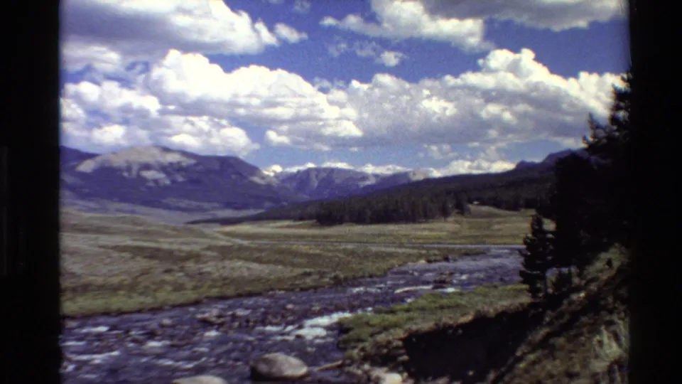 1980 home-movie frame of a river curving past grassland toward snow-streaked mountains, with dark trees and film border at the right edge.