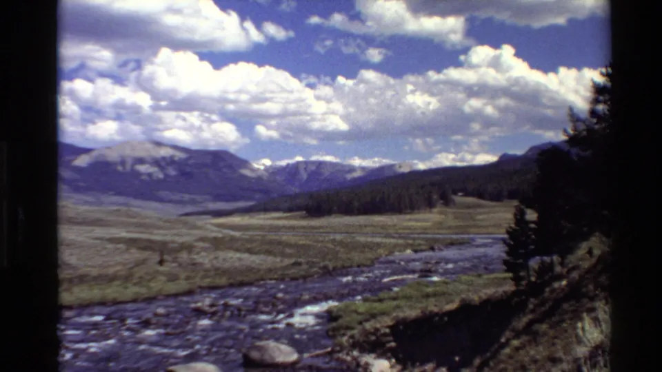 8mm Western USA landscape showing grass plains, a rocky river channel, dark tree line, distant mountains, and large white clouds.