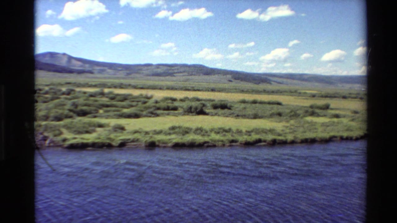 1980 Western USA 8mm frame showing a broad river in front of green meadows, low hills, distant mountains, and scattered clouds.