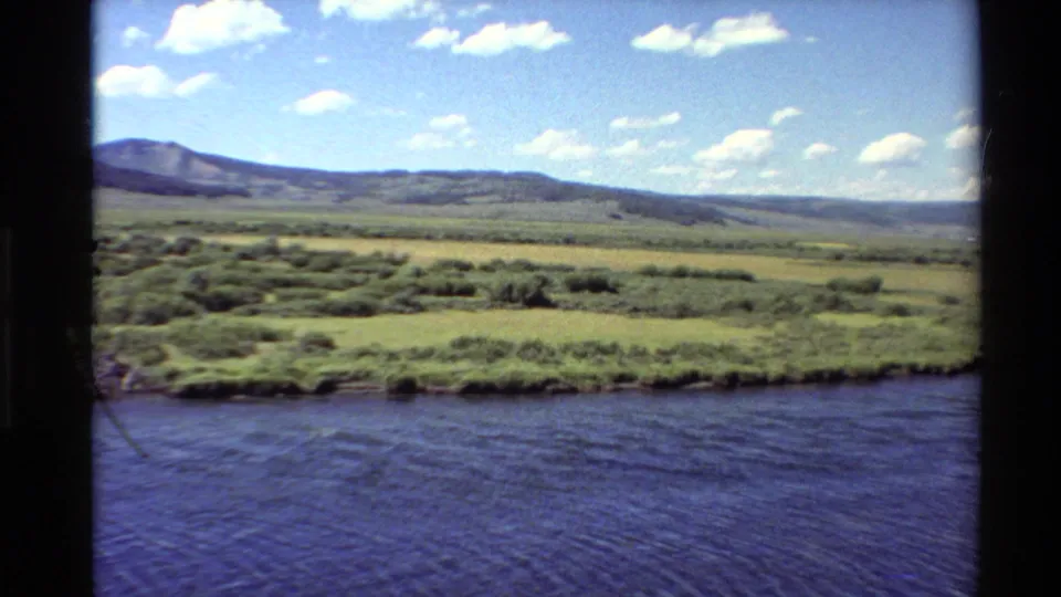 1980 Western USA 8mm frame showing a broad river in front of green meadows, low hills, distant mountains, and scattered clouds.