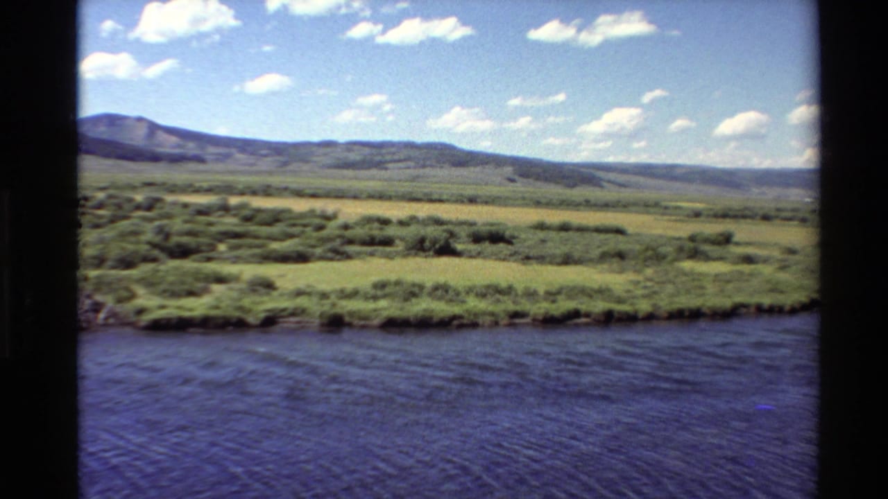 River foreground, green meadow, hills, mountains, and scattered clouds.