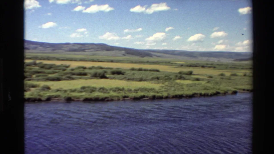 Western USA 1980 8mm landscape frame showing rippled river water, grassy meadow, shrub patches, and hazy hills beneath clouds.