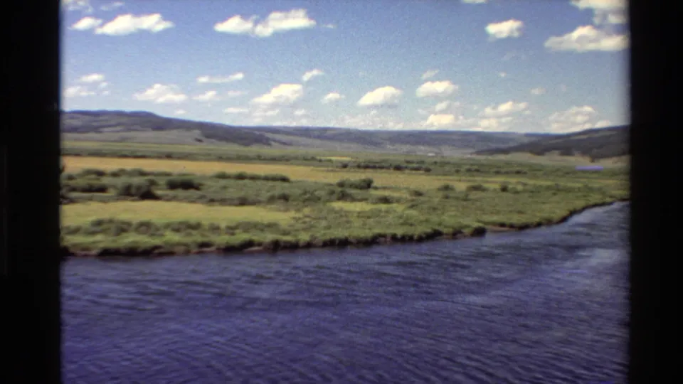 1980 Western USA 8mm frame showing the river curving along a grassy bank, with meadows, low hills, and scattered clouds in view.