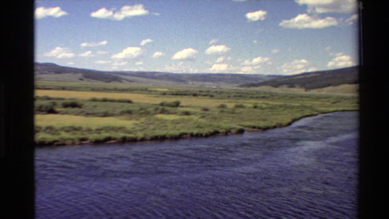 1980 Western U.S. 8mm view of a broad river or lake edge, green valley, low hills, and scattered clouds.