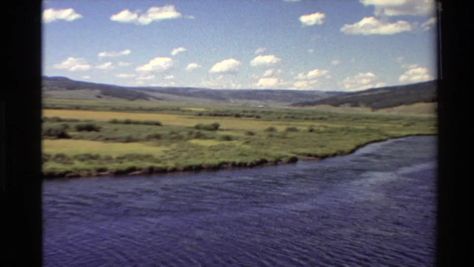 1980 Western U.S. 8mm view of a broad river or lake edge, green valley, low hills, and scattered clouds.