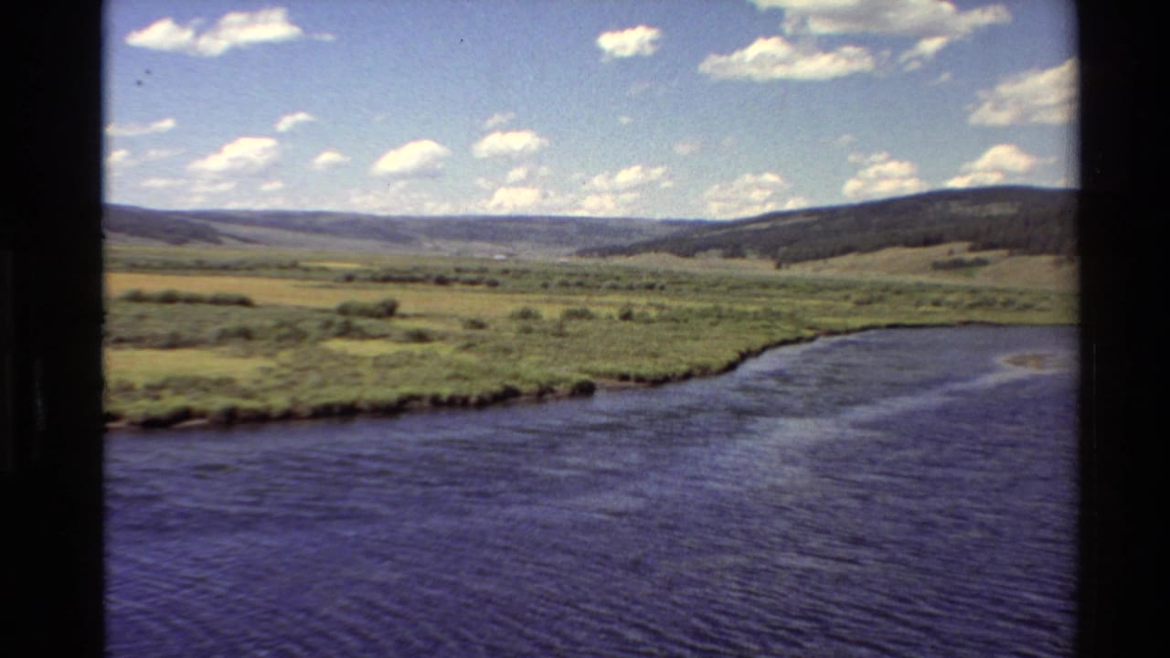 8mm Western U.S. landscape with rippled water, green meadow, rolling hills, and scattered clouds in the distance.
